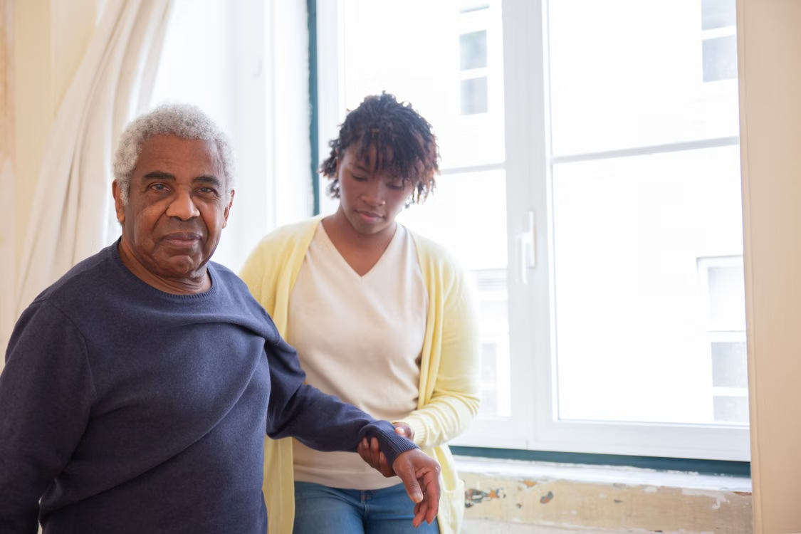 A young caregiver helping an elderly man adjust his shirt.